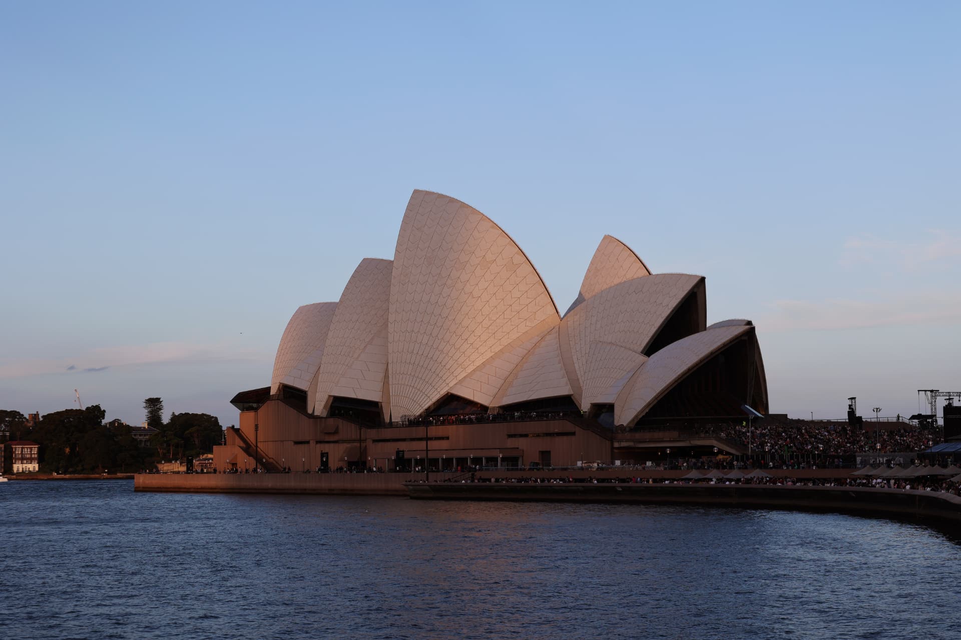 The Sydney Opera House at dusk, its white sails glowing above the harbour with crowds along the forecourt