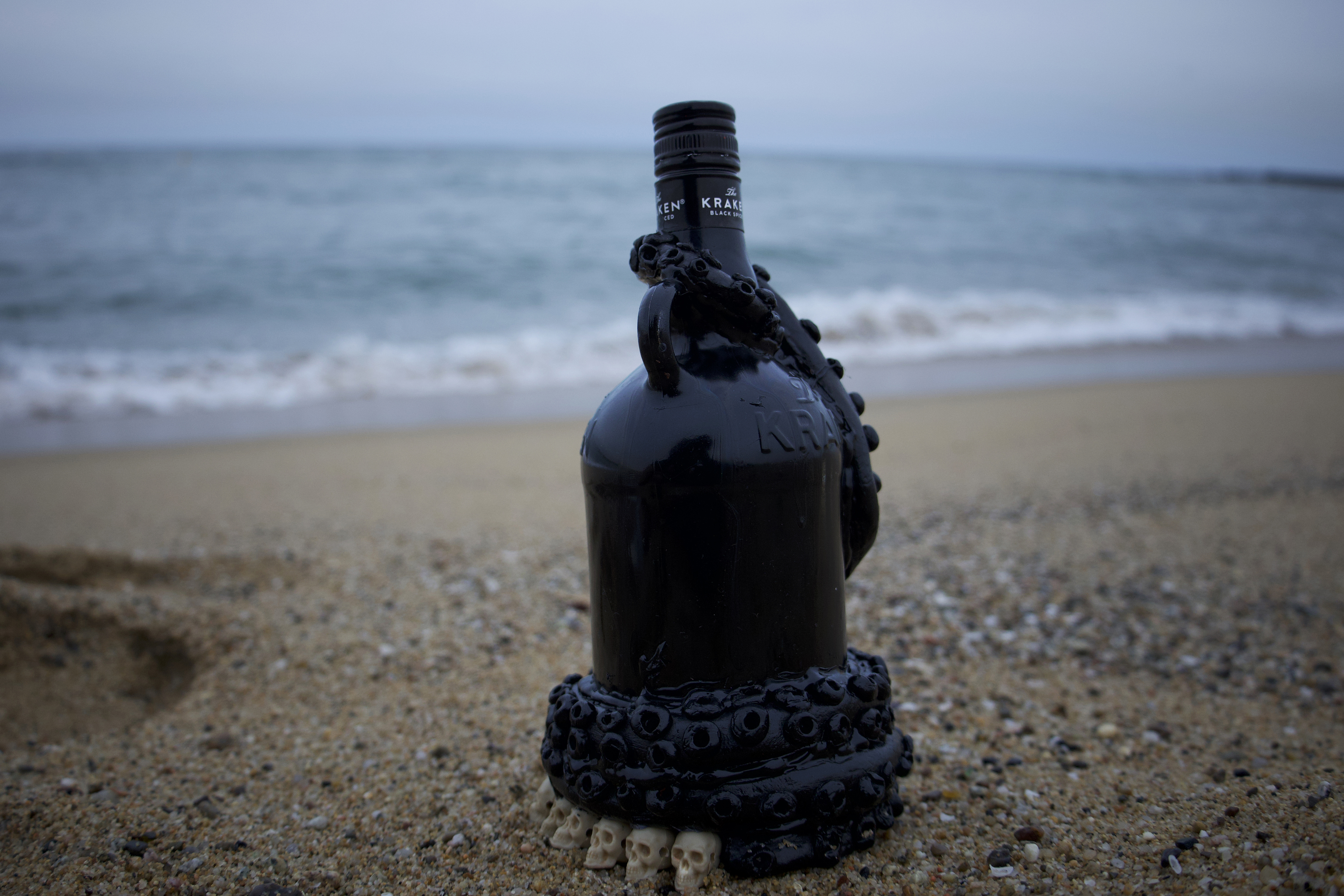 A black Kraken rum bottle decorated with sculpted tentacles and small skulls, standing on a beach with the sea behind it.