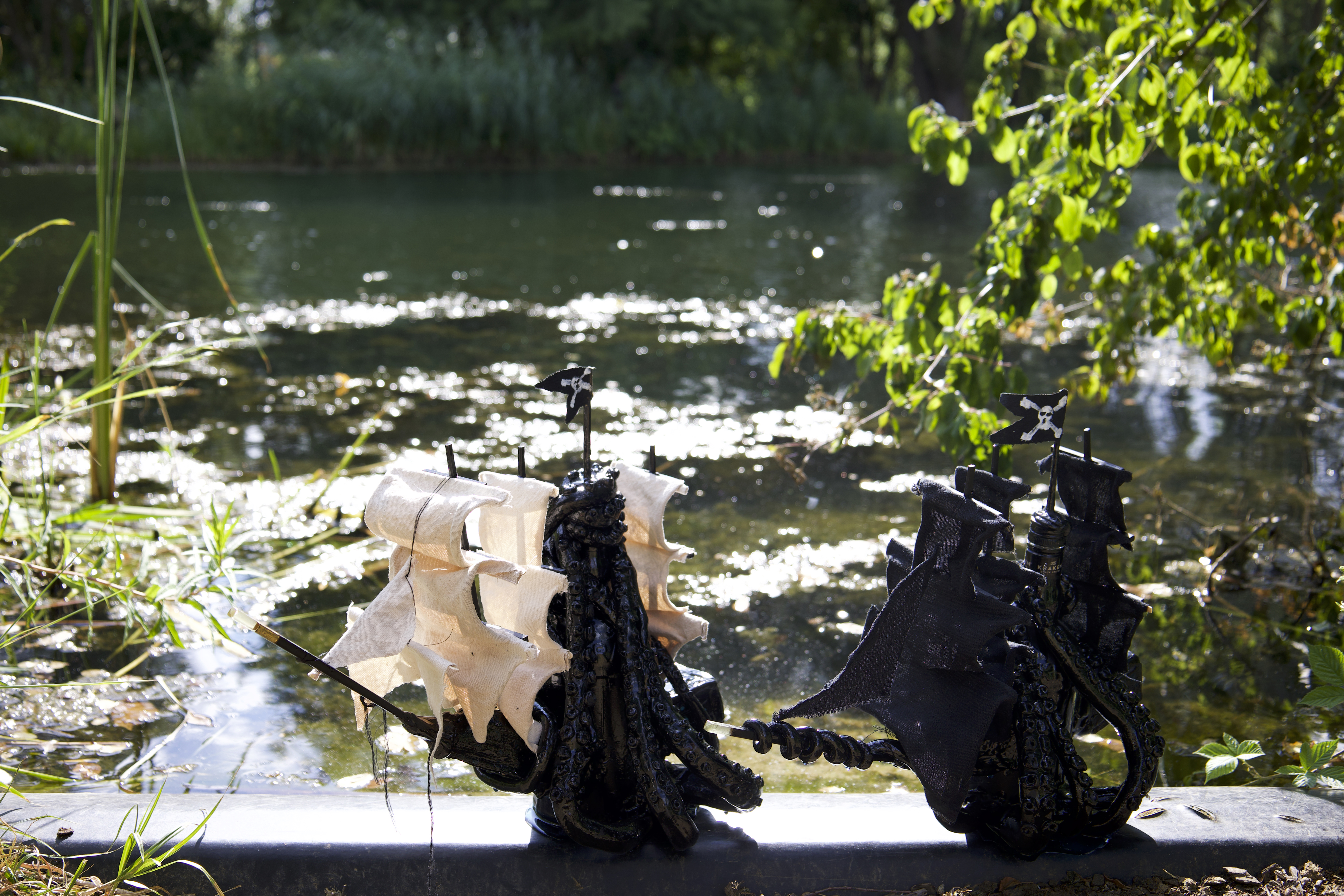 Two pirate-ship sculptures made from Kraken rum bottles, one with cream sails and one with black sails, beside a pond.