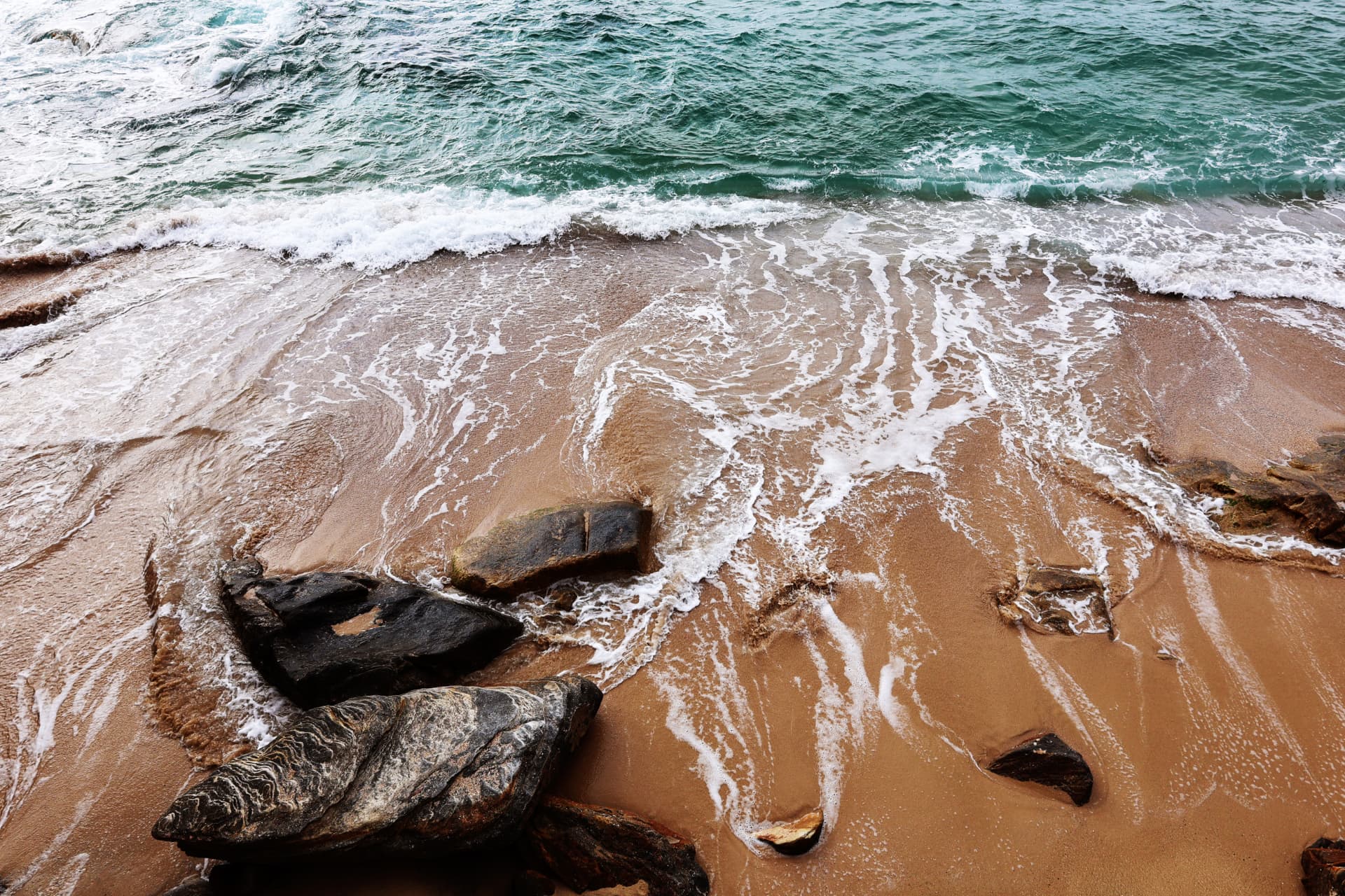 Turquoise waves washing over golden sand and dark rocks along the shoreline at Watsons Bay, NSW, Australia