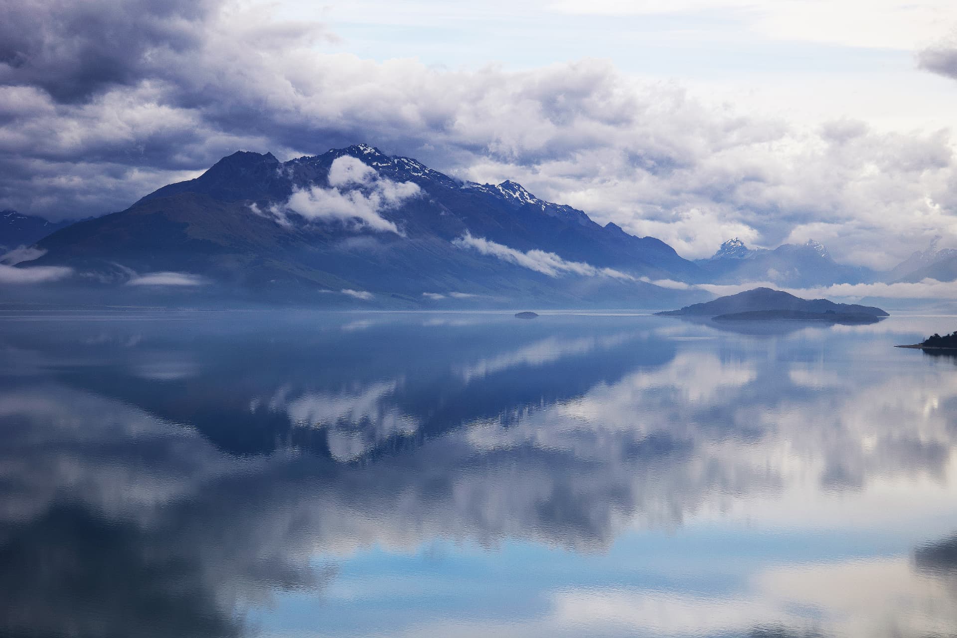 A calm lake reflecting cloud-covered mountains and islands in the Otago Region of New Zealand