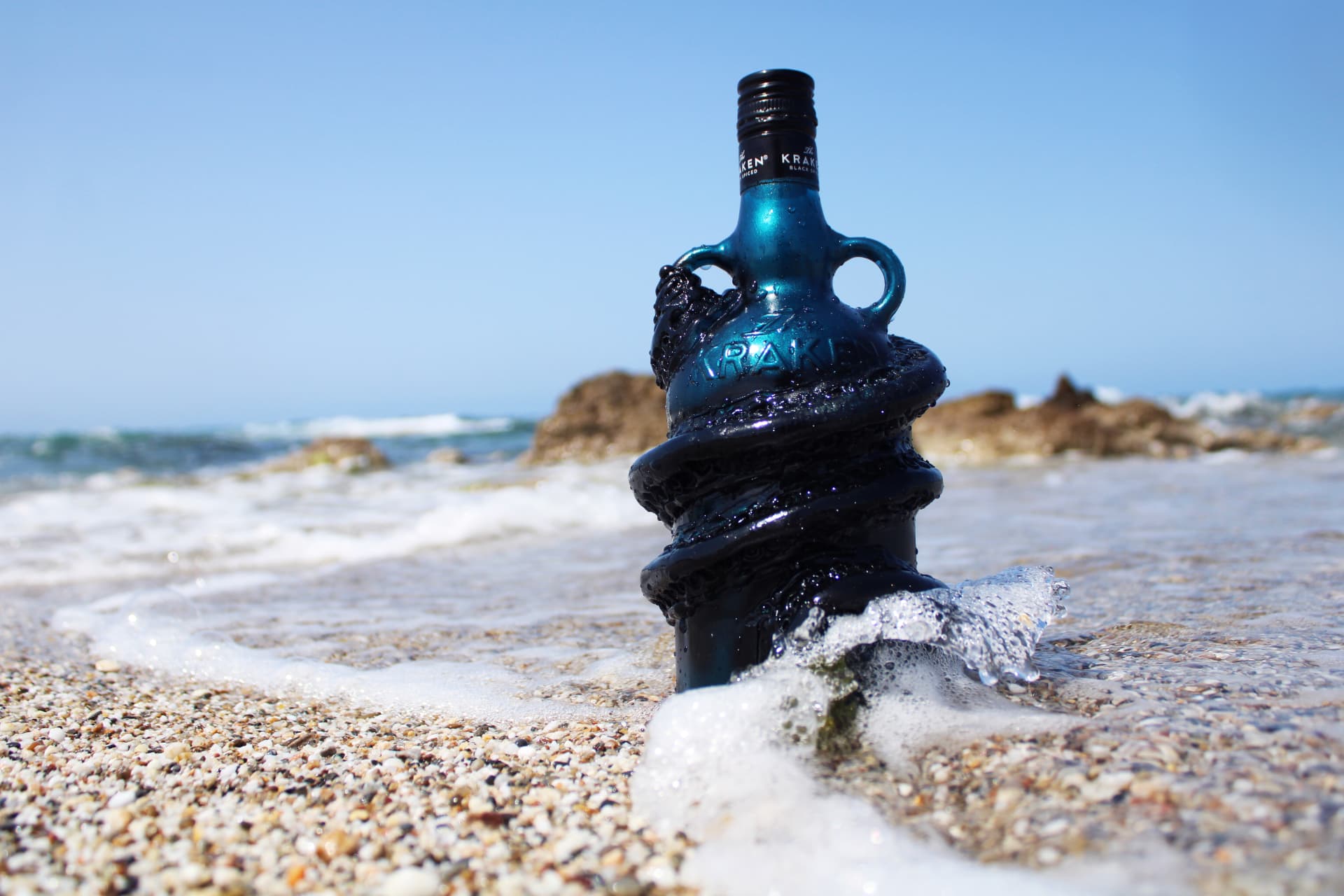 A blue Kraken rum bottle wrapped with sculpted black tentacles, standing upright in surf on a sunny pebble beach.