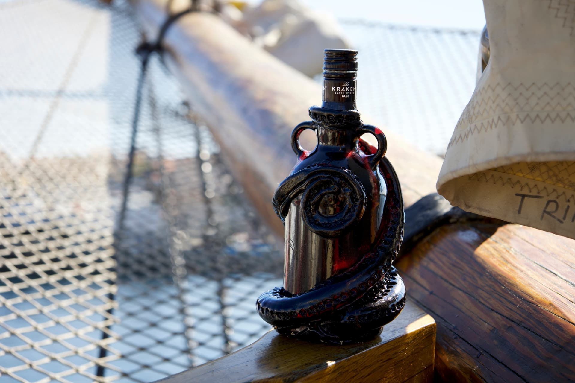 A black Kraken rum bottle sculpted with coiled tentacles, resting on a wooden boat deck beside a sail.