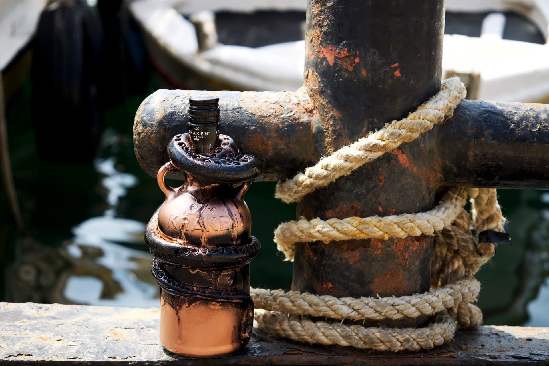 A copper-colored Kraken rum bottle sculpted with dark tentacles and aged patina, tied to a weathered black pier post wrapped with thick rope.