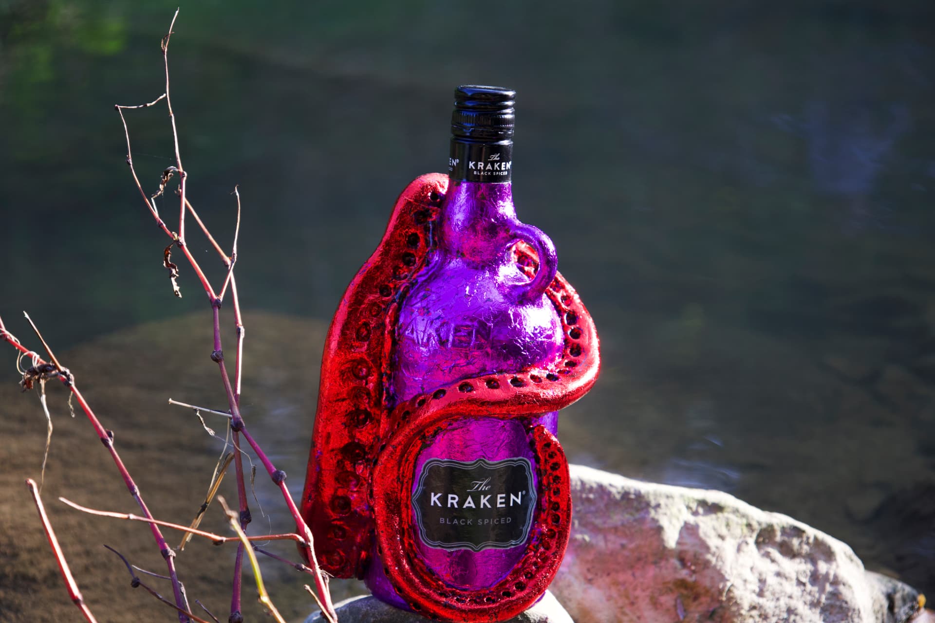 A purple Kraken rum bottle wrapped with sculpted red tentacles, standing on a rock beside calm water with bare branches in the foreground.