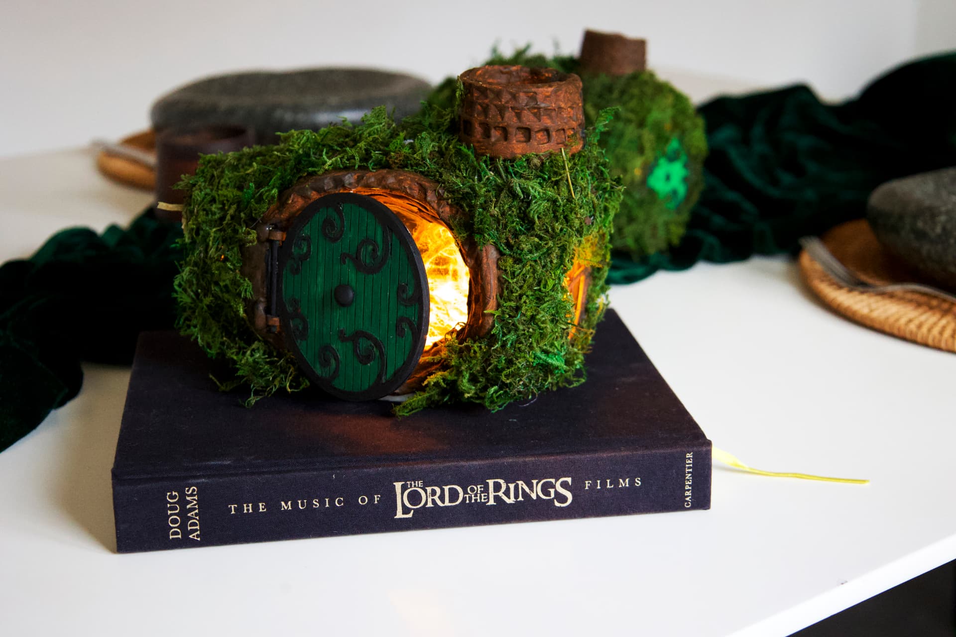 A handmade Hobbit house sculpture with a green round door and warm interior light, nestled in moss and greenery.