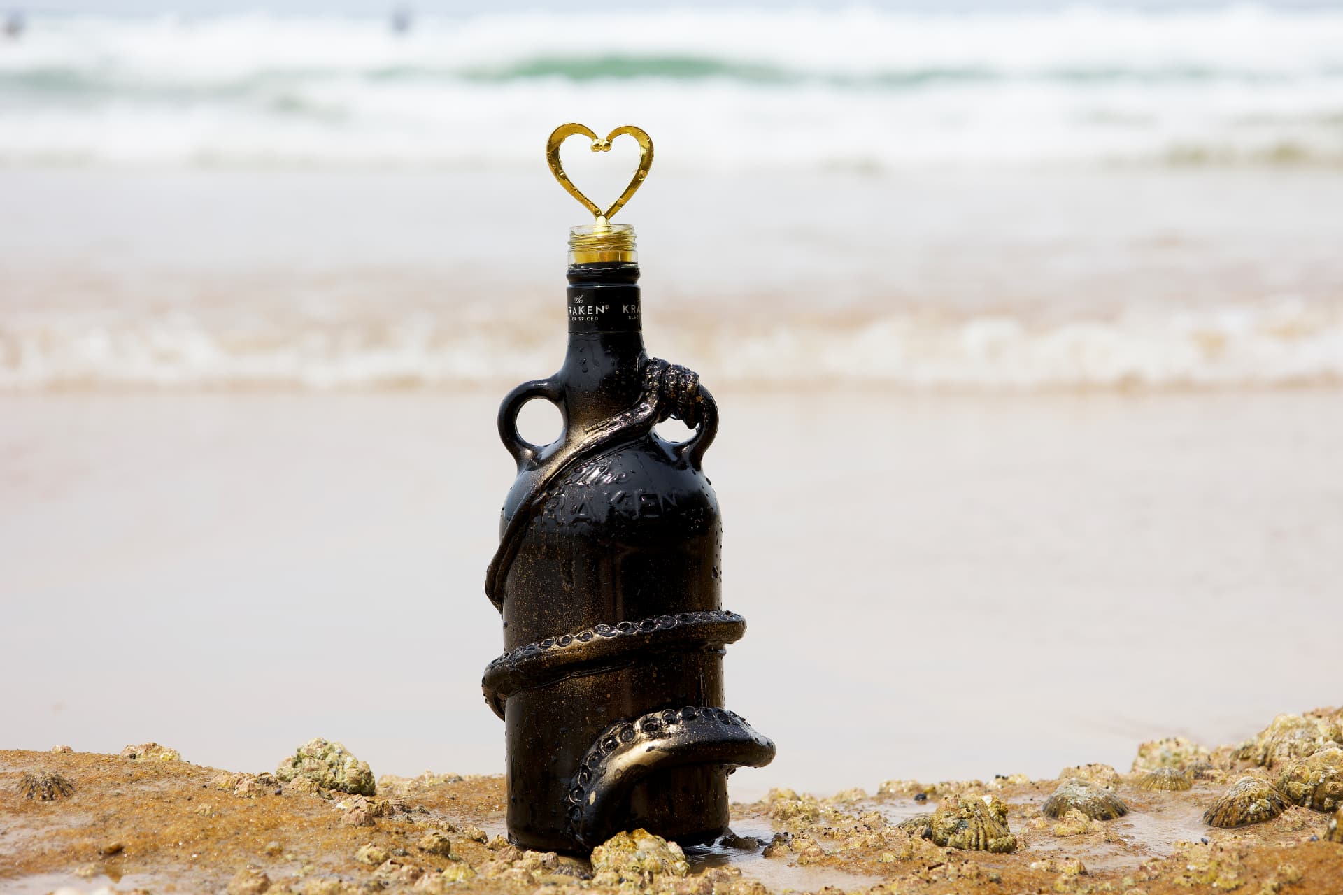 A black Kraken rum bottle decorated with sculpted tentacles and a golden heart-shaped wire, standing on a sandy beach with gentle surf behind it.