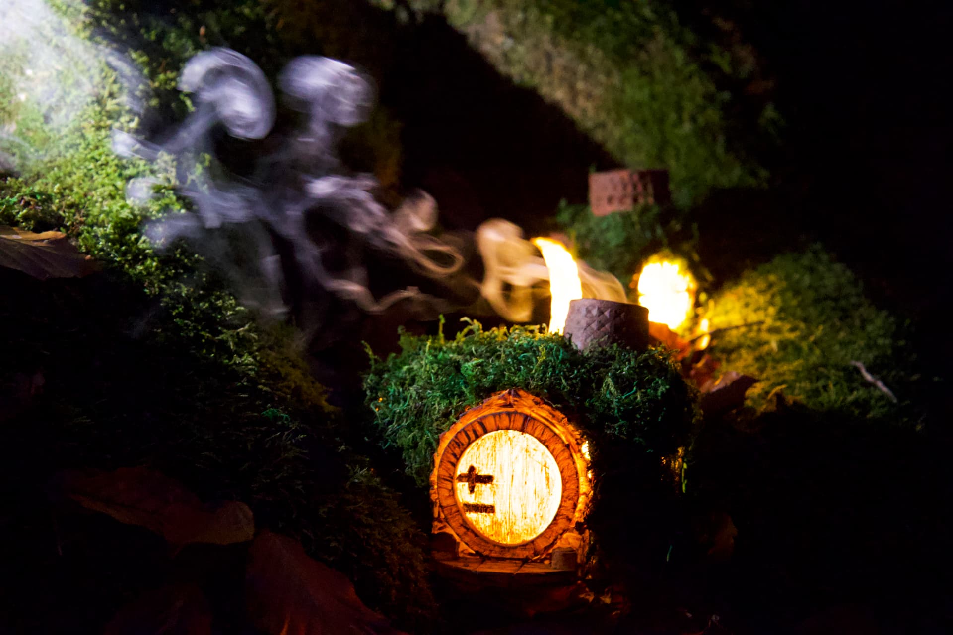 A handmade Hobbit house sculpture with a round wooden door, set in moss with candlelight and smoke at night.