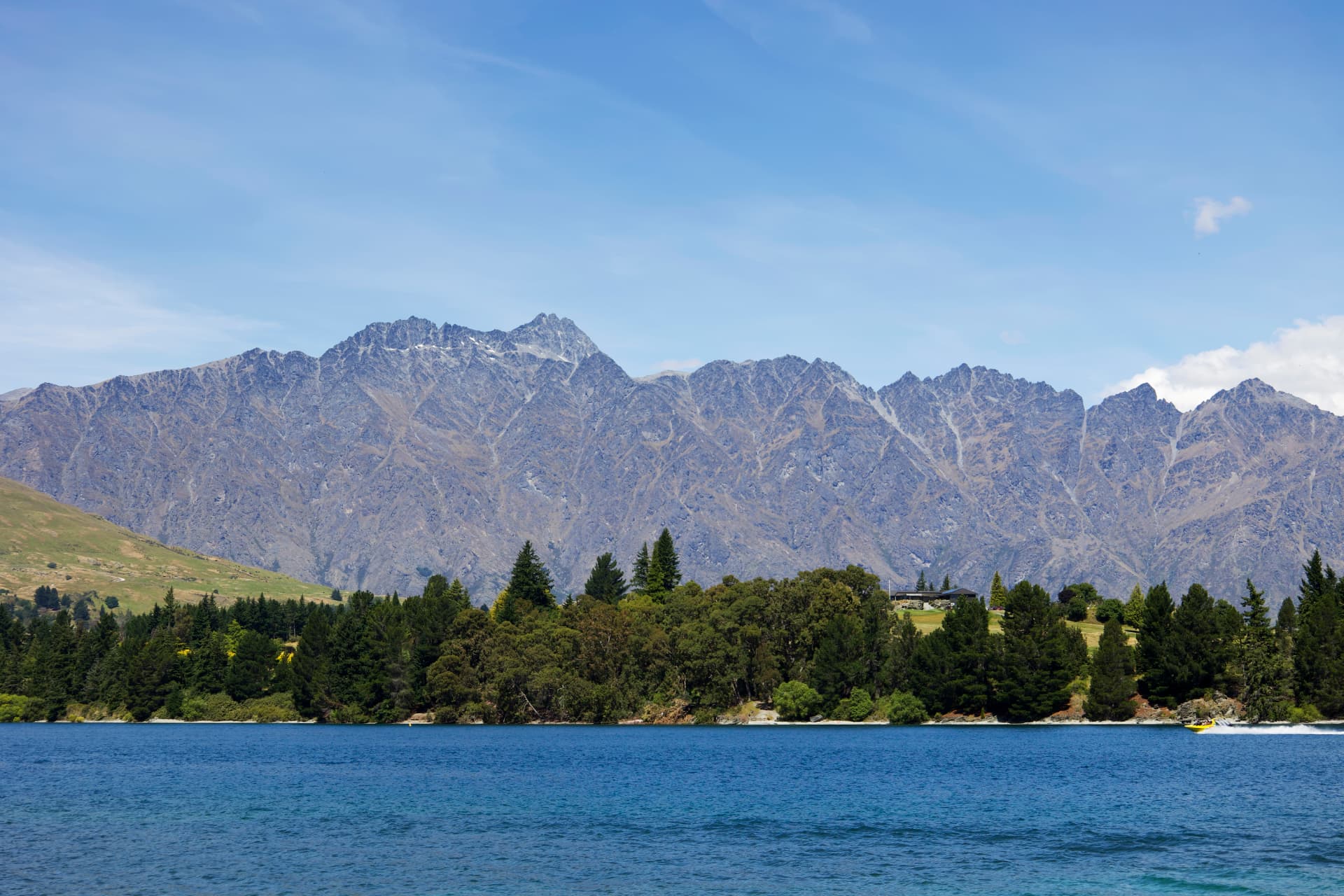 The Remarkables mountain range rising above Lake Wakatipu near Queenstown, with a small boat on the bright blue water