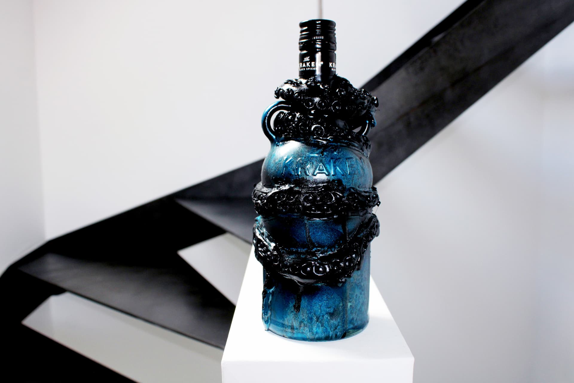 A deep blue Kraken rum bottle heavily coated with sculpted black tentacles and barnacles, standing on a white pedestal against a modern staircase.