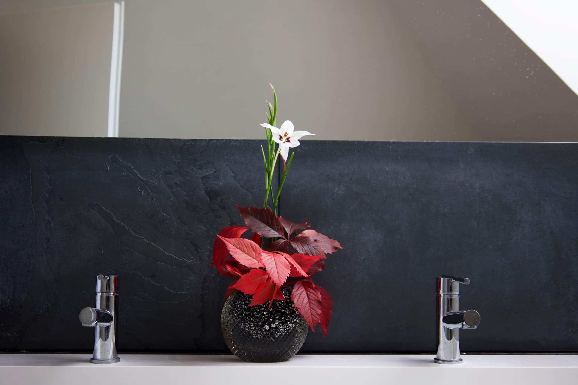 An ikebana arrangement with red leaves and a white lily in a dark stone vase, set against a slate wall.