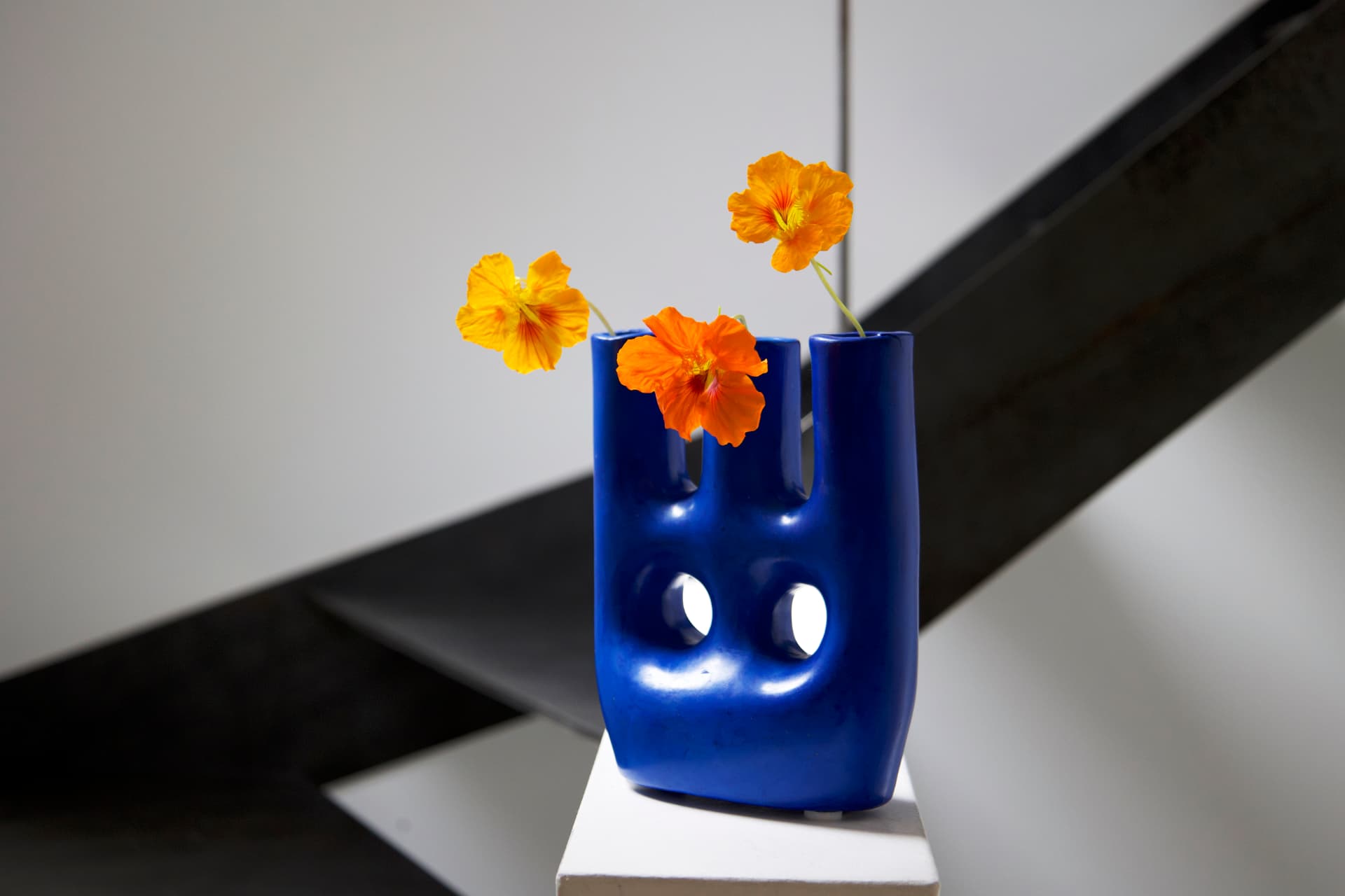 A sculptural blue ceramic vase with orange nasturtium flowers, displayed on a white pedestal.