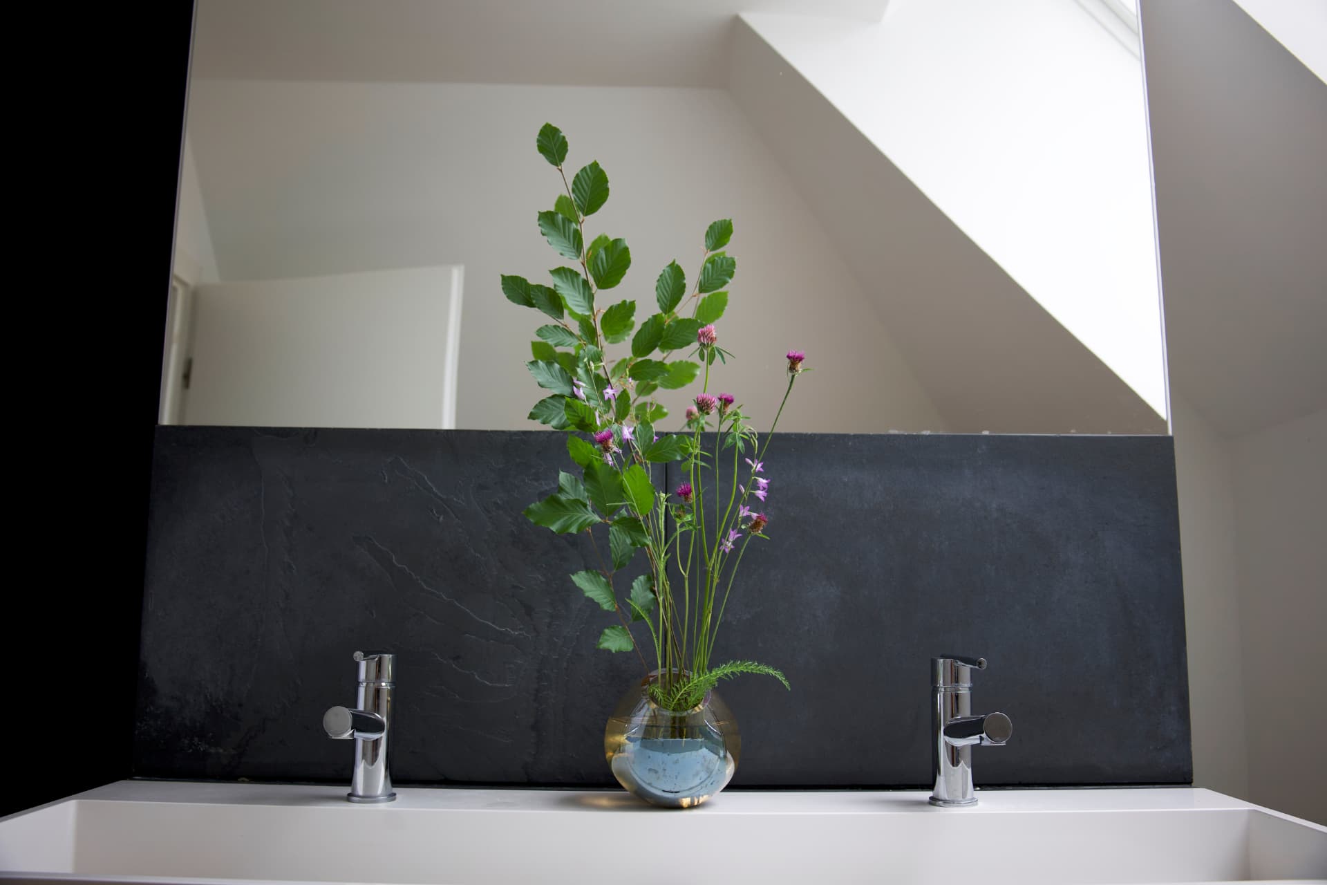 An ikebana arrangement with green beech branches and purple clover in a round glass vase.