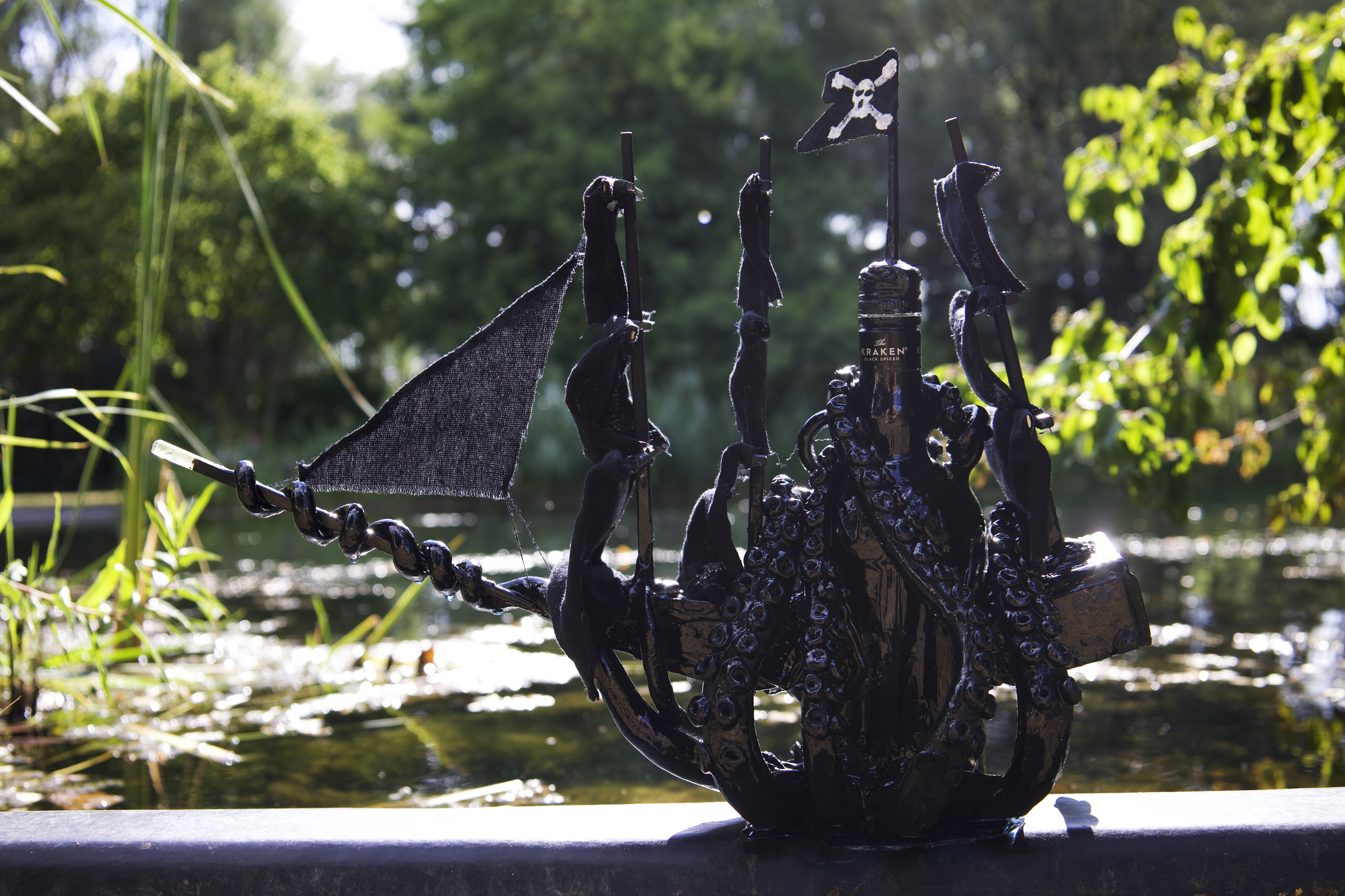 A single black-sailed pirate ship sculpture made from a Kraken rum bottle, outdoors beside a pond.