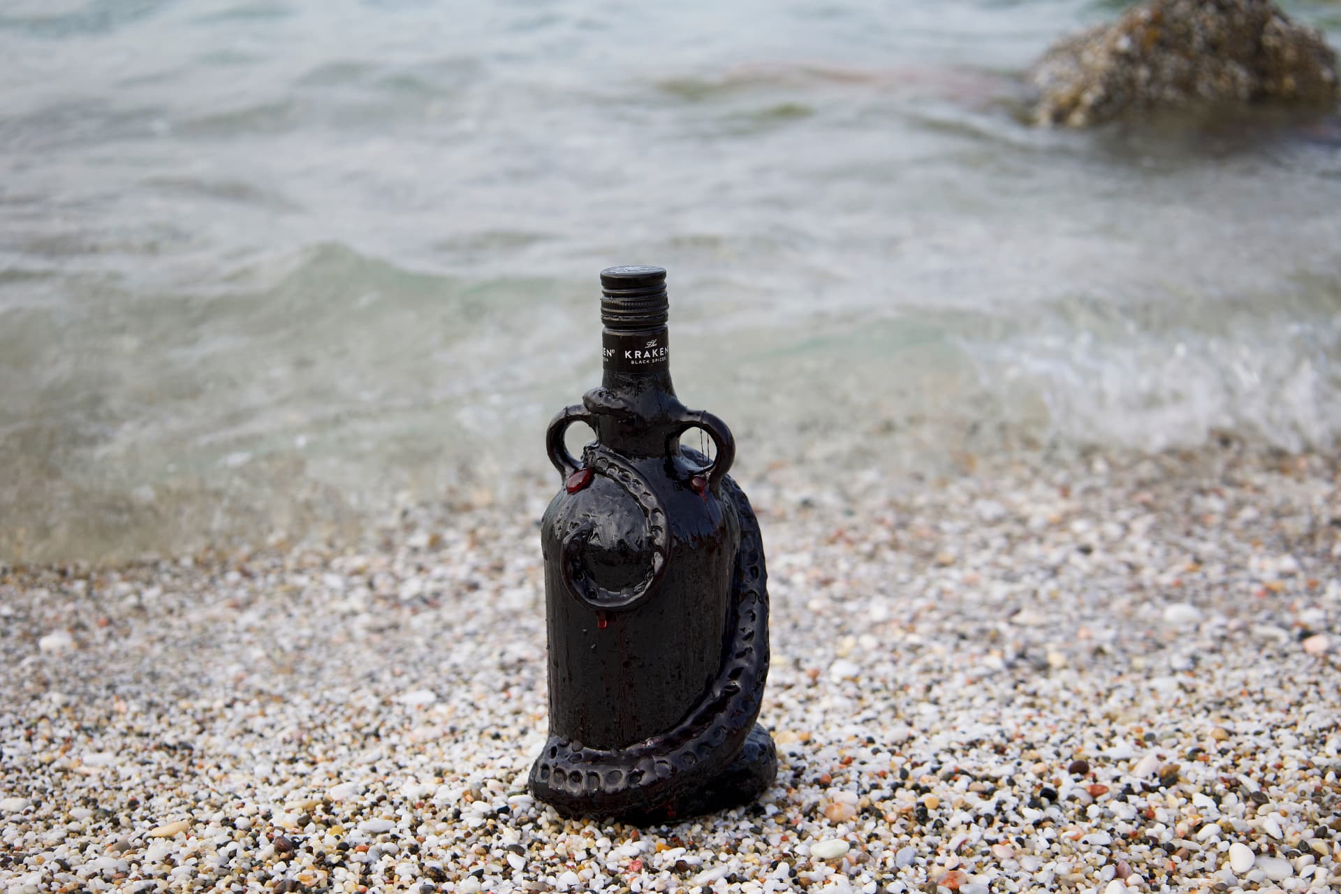 A dark Kraken rum bottle with sculpted black tentacles and red accents, standing on a pebble beach at the water's edge.
