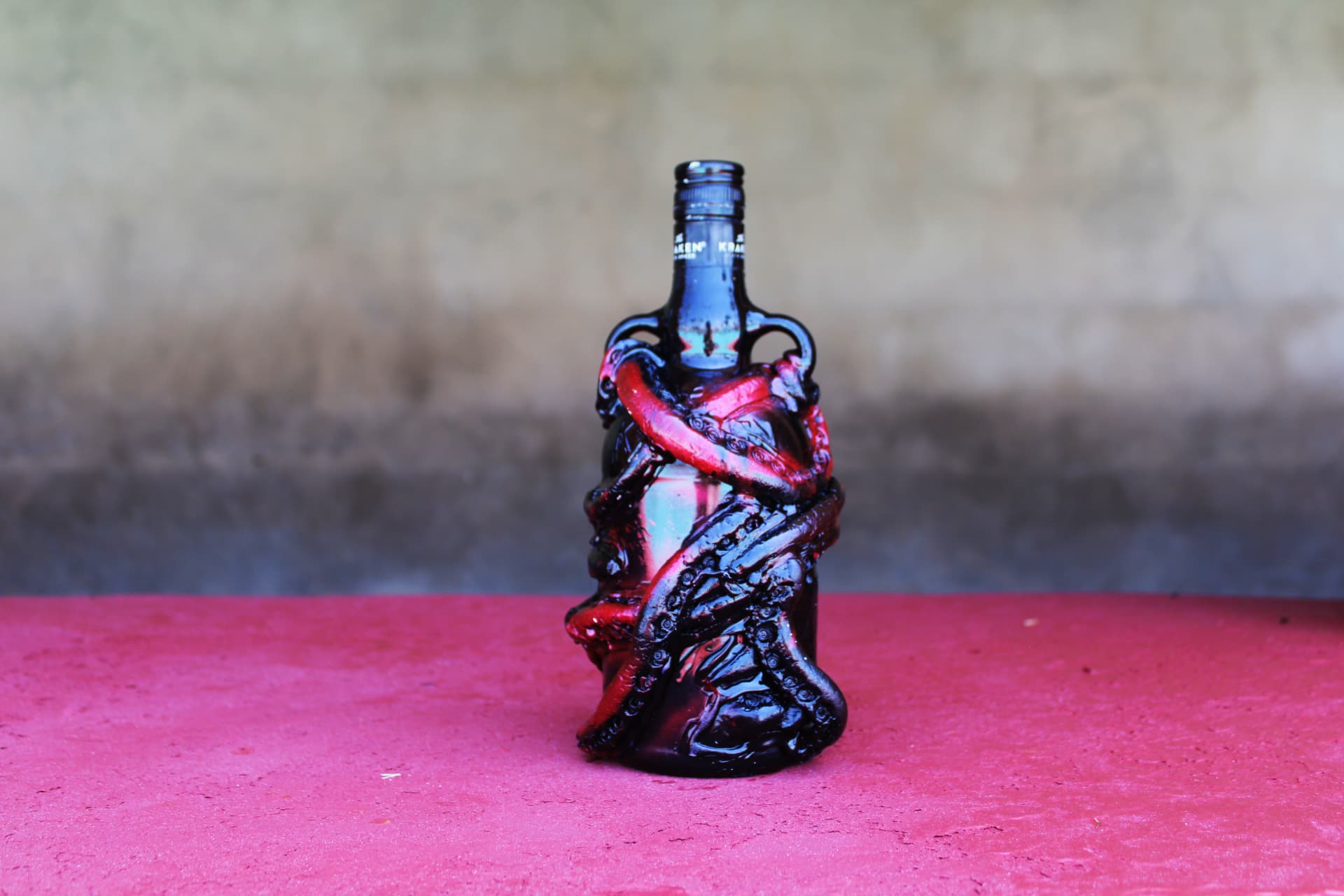A black Kraken rum bottle sculpted with dark and red tentacles, standing on a bright pink surface against a weathered wall.