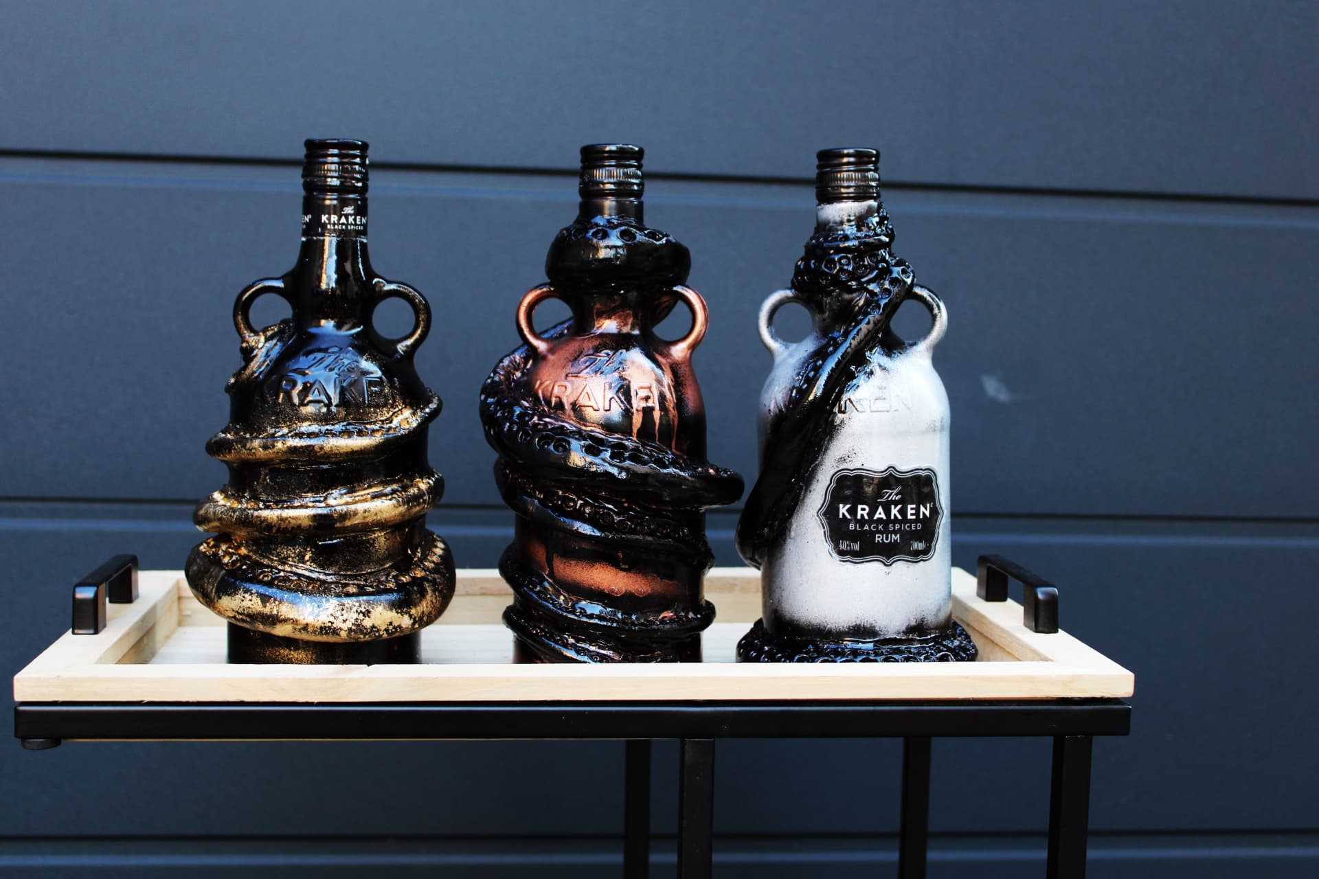 Three sculpted Kraken rum bottles in black, copper, and white finishes, arranged on a wooden tray against a dark background.