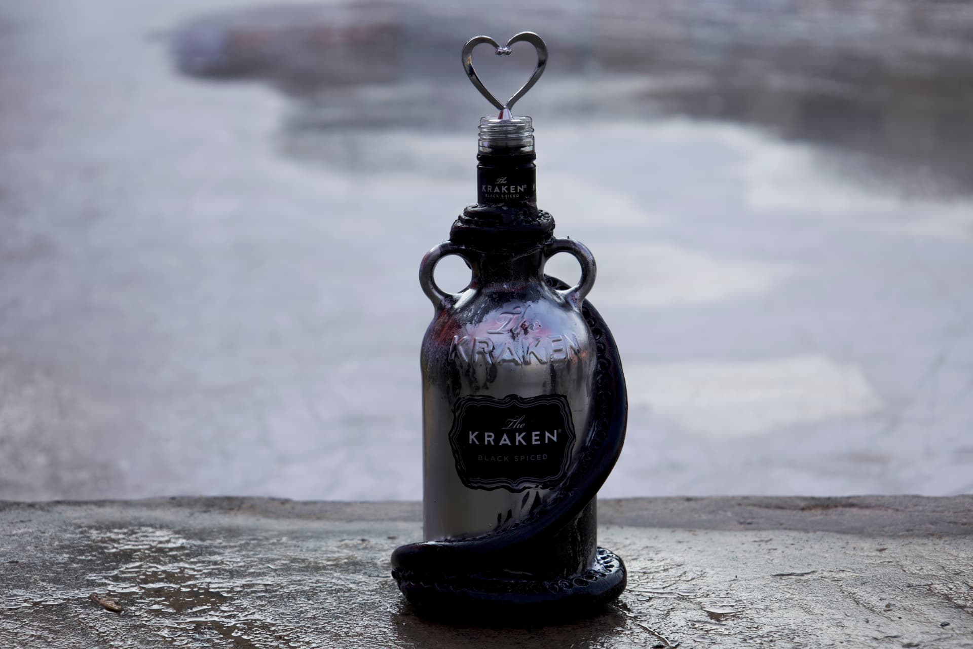 A metallic silver Kraken rum bottle with sculpted black tentacles and a heart-shaped bottle stopper, standing on a wet stone surface.